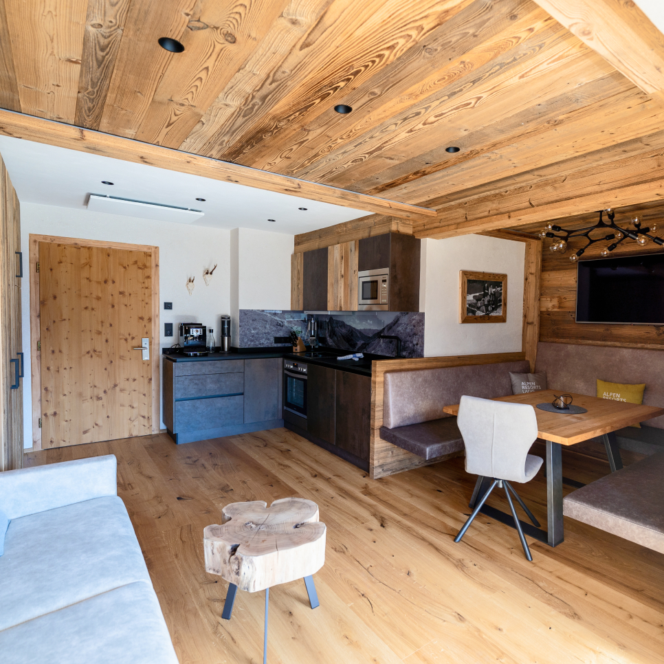Eine gemütliche Holzhütte mit Küche und Essbereich. Die Küche verfügt über dunkle Holzschränke und moderne Geräte. Die Essecke umfasst einen Holztisch mit gepolsterten Sitzgelegenheiten. Sichtbare Holzbalken und ein kleiner Holztisch verleihen rustikalen Charme.