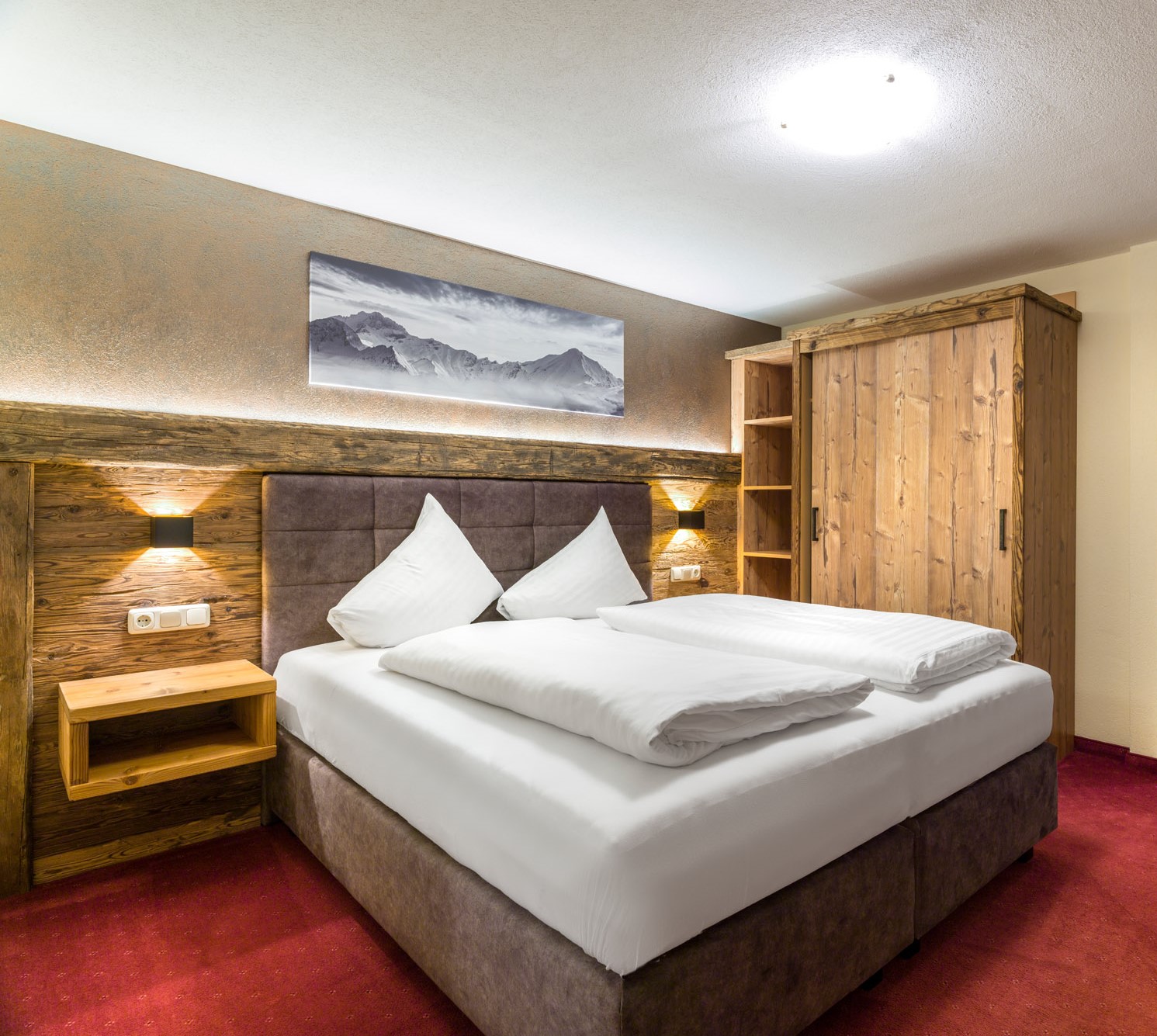 Ein gemütliches Apartment für 2-3 Personen in Ladis erwartet Sie mit einem sauber gemachten Doppelbett, einem hölzernen Kopfteil und Nachttischen. Über dem Bett hängt ein Bild einer Berglandschaft. Rechts davon steht ein Holzschrank. Das Zimmer ist mit sanfter Beleuchtung und rotem Teppich ausgestattet.