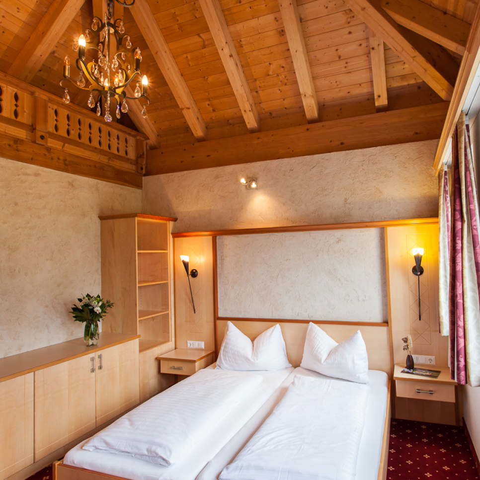 Ein gemütliches Schlafzimmer im Apartment Abendsonne im Alpenapart in Ladis verfügt über eine Holzdecke und einen Kronleuchter. Dieses rustikale Zimmer verfügt über ein Doppelbett mit weißer Bettwäsche, zwei Wandlampen, einen Blumenstrauß auf dem Schrank und gemusterte Vorhänge, die seinen warmen Charme unterstreichen.