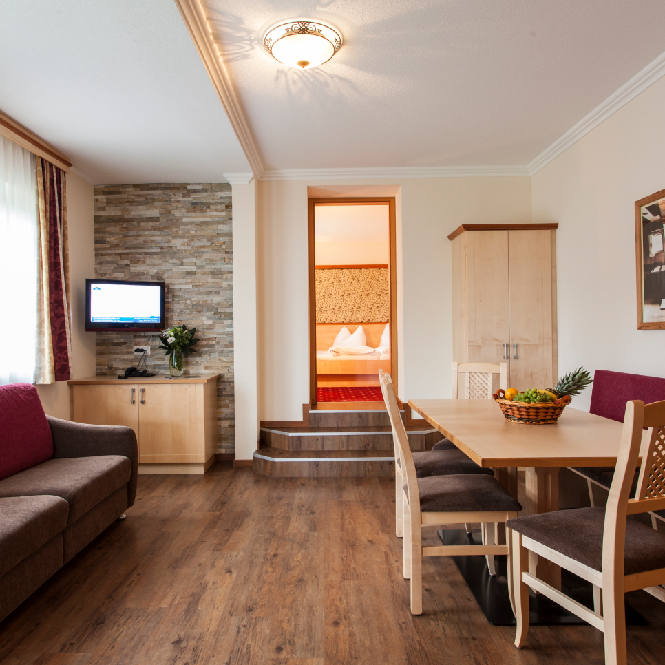 Erleben Sie den entspannten Charme dieses Alpenapart-Apartments für 4-6 Personen. Es verfügt über ein gemütliches Wohnzimmer mit Holzboden, einem braunen Sofa mit violetten Kissen und einem Esstisch mit einem Obstkorb. Ein Fernseher ist an einer Steinwand angebracht und öffnet sich zu einem warm beleuchteten Schlafzimmer.