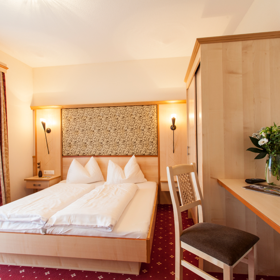 Gemütliches Hotelzimmer, perfekt für ein Paar, mit einem sauber gemachten Doppelbett mit weißer Bettwäsche und Holzmöbeln. Ein gemustertes Kopfteil ergänzt das Dekor, während ein kleiner Schreibtisch und Stuhl die Funktionalität erhöhen. Genau wie in einem Apartment für 4-6 Personen im Alpenapart in Ladis sorgt warmes Licht für eine einladende Atmosphäre.