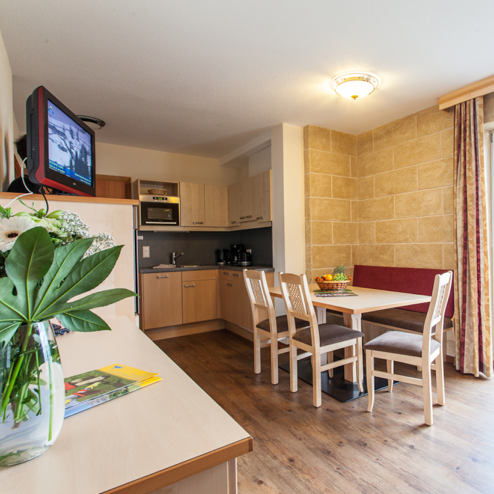 Eine gemütliche Küche mit hellen Holzmöbeln empfängt die Gäste in diesem charmanten Alpenapart in Ladis. Sie ist perfekt für ein Apartment für 4-6 Personen und verfügt über einen kleinen Esstisch, einen an der Wand montierten Fernseher und eine mit Blumen und Zeitschriften geschmückte Arbeitsplatte. Eine Obstschale ziert den Holztisch.