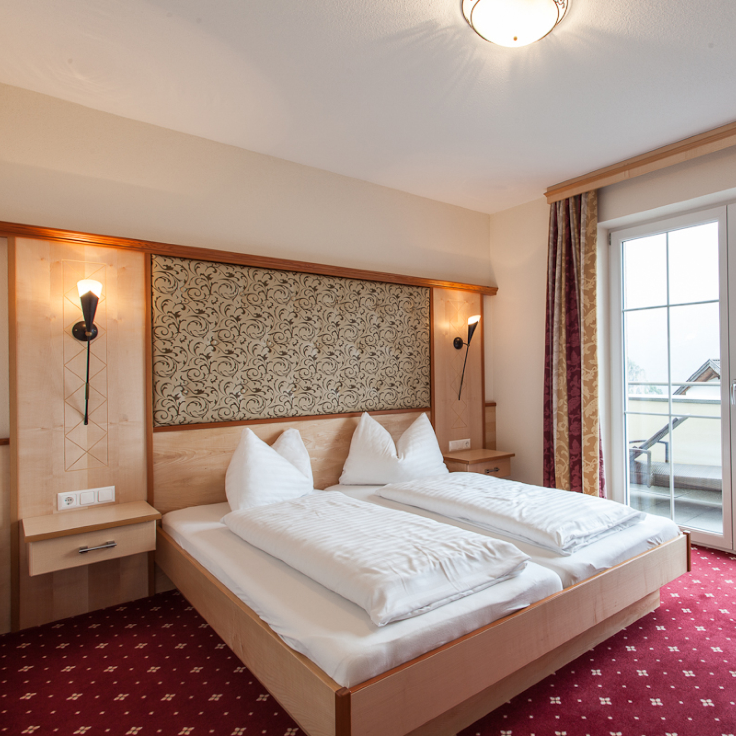 Ein ordentliches Hotelzimmer im charmanten Alpenapart in Ladis verfügt über ein Doppelbett aus Holz mit weißer Bettwäsche und Kissen. Das Kopfteil ist kunstvoll gestaltet, während zwei Wandlampen passende Nachttische beleuchten. Ein Fenster mit teilweise zugezogenen Vorhängen verleiht eine ruhige Note, perfekt für ein Apartment für 4 Personen.