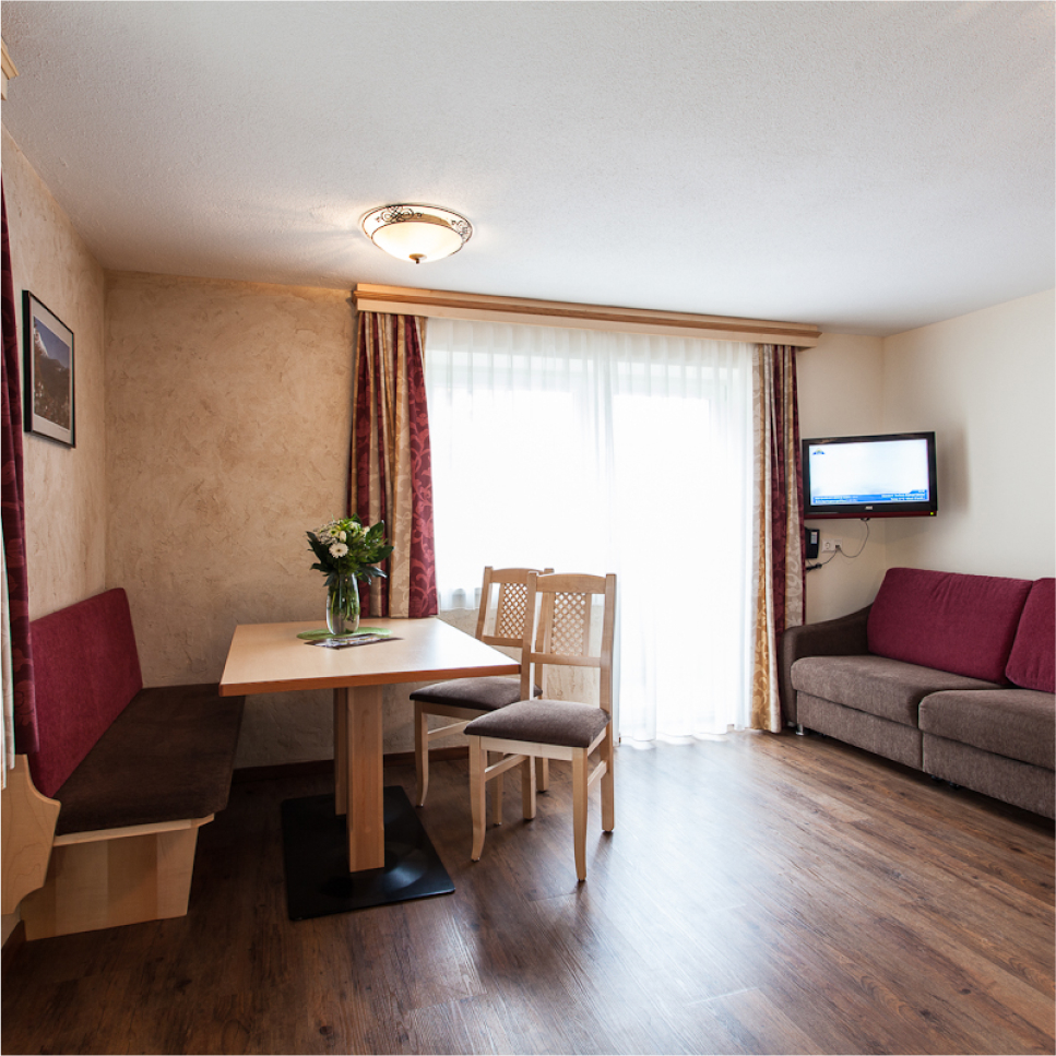 Ein gemütliches Apartment für 2-4 Personen im Alpenapart in Ladis, mit einem Essbereich mit einem Holztisch, zwei Stühlen und einer Bank mit roten Kissen. Ein kleiner Blumenstrauß schmückt den Tisch. Ein bequemes Sofa und ein an der Wand montierter Fernseher befinden sich in der Nähe, während große Fenster mit transparenten Vorhängen den Raum mit natürlichem Licht füllen.