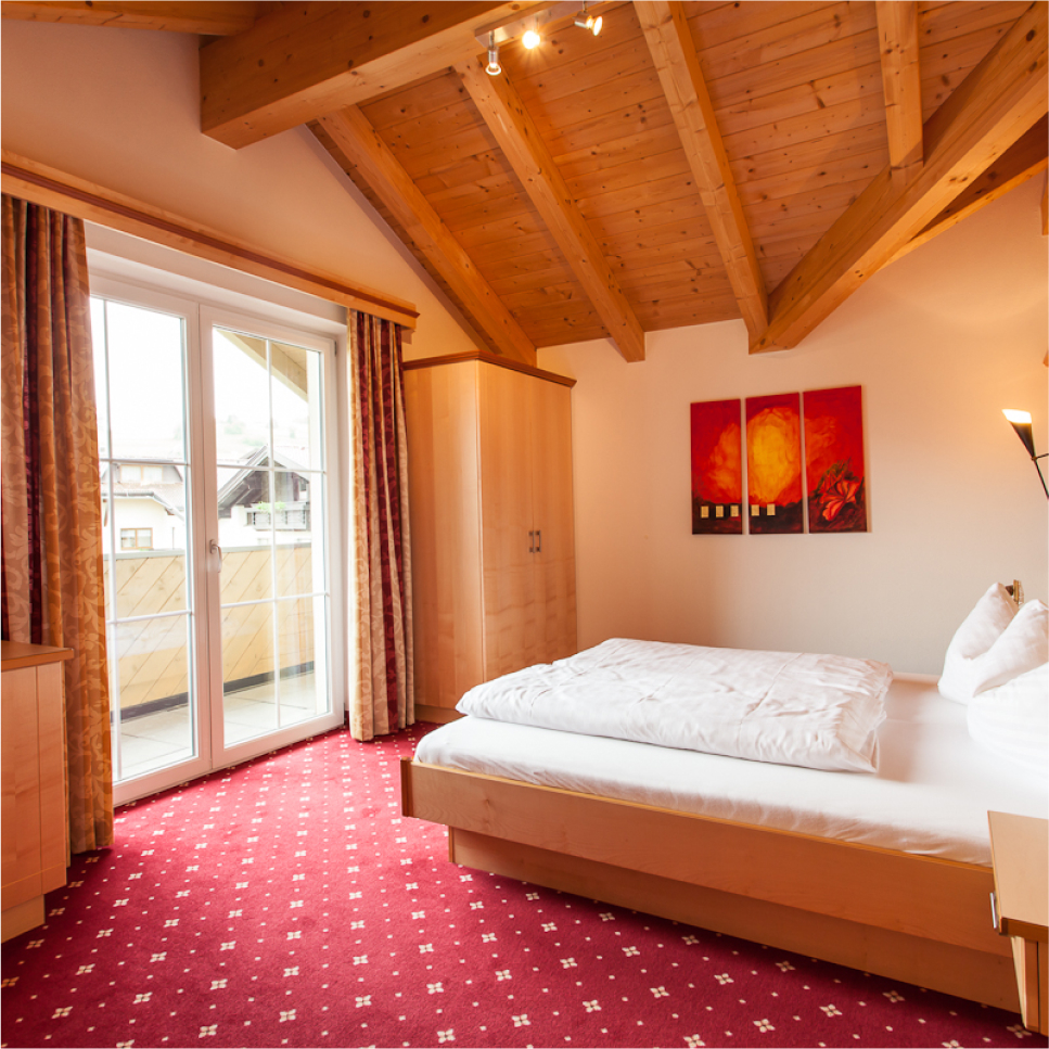 Dieses gemütliche Schlafzimmer im Apartment Morgensonne für 2-4 Personen in Ladis verfügt über eine Holzdecke und einen roten Teppich. Es verfügt über ein ordentlich gemachtes Doppelbett, Holzmöbel und farbenfrohe Wandkunst. Große Fenster mit Vorhängen öffnen sich zu einem Balkon und durchfluten den Raum mit natürlichem Licht.
