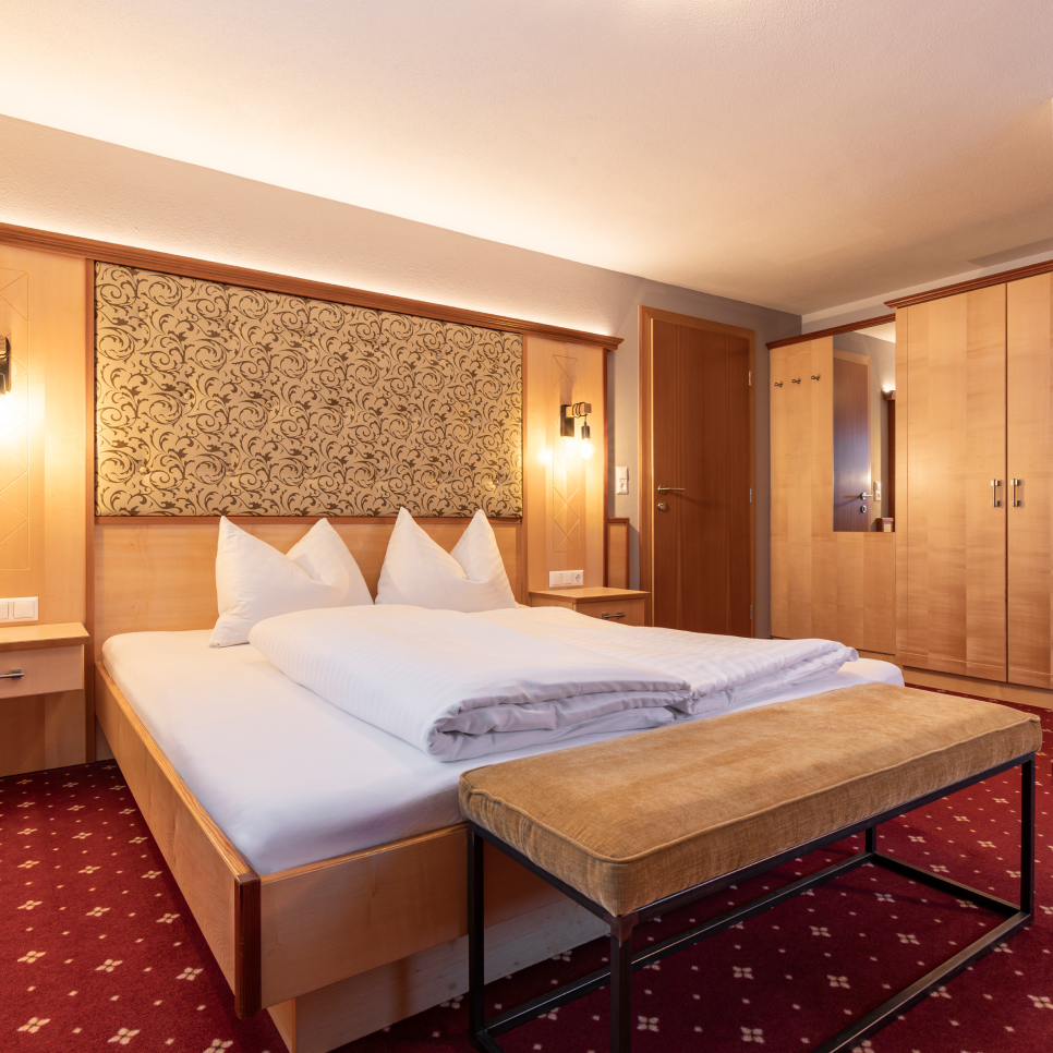 Ein gemütliches Hotelzimmer, perfekt für Urlaub zu Zweit in Serfaus-Fiss-Ladis, mit einem Doppelbett mit weißer Bettwäsche und Kissen. Es verfügt über ein hölzernes Kopfteil mit kunstvollem Design, eine Bank am Fußende, einen hölzernen Kleiderschrank und Nachttische. Der rote Teppich hat ein dezentes Muster für zusätzliche Eleganz.