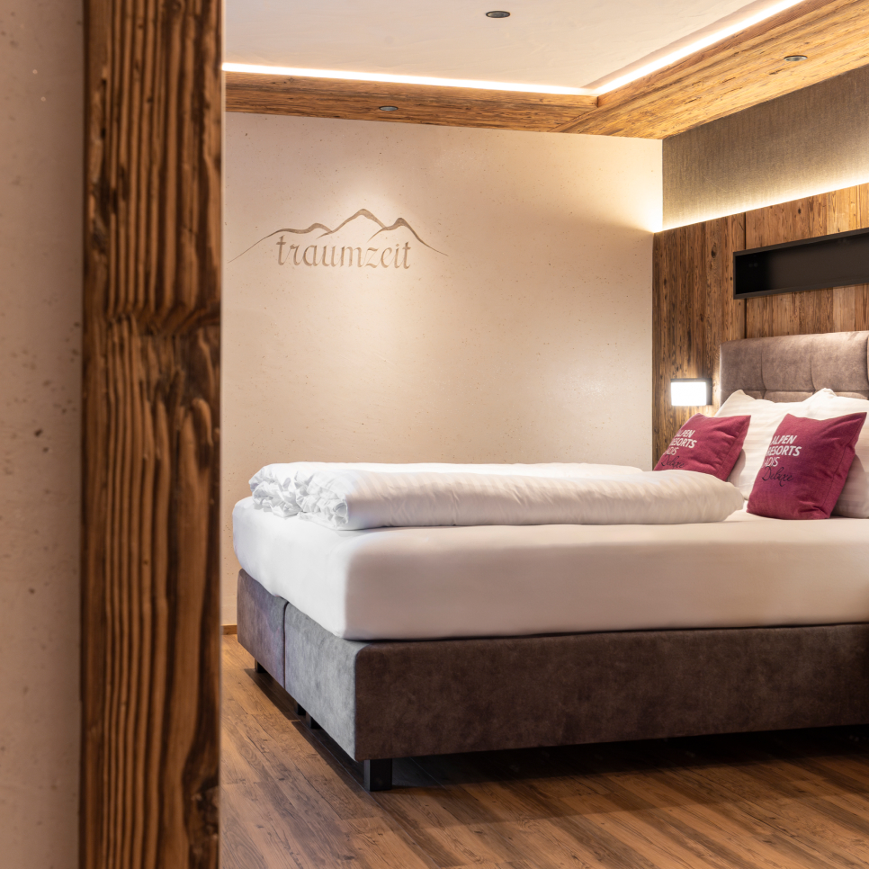 Ein gemütliches Alpin-Chalet-Hotelzimmer in Ladis verfügt über ein weiches, ordentlich gemachtes Bett mit weißer Bettwäsche und roten Kissen. Holzwände und -böden sorgen für Wärme, während „traumzeit“ die Wand darüber elegant schmückt. Sanfte Beleuchtung rundet diesen perfekten Rückzugsort für große Gruppen ab, die Entspannung suchen.