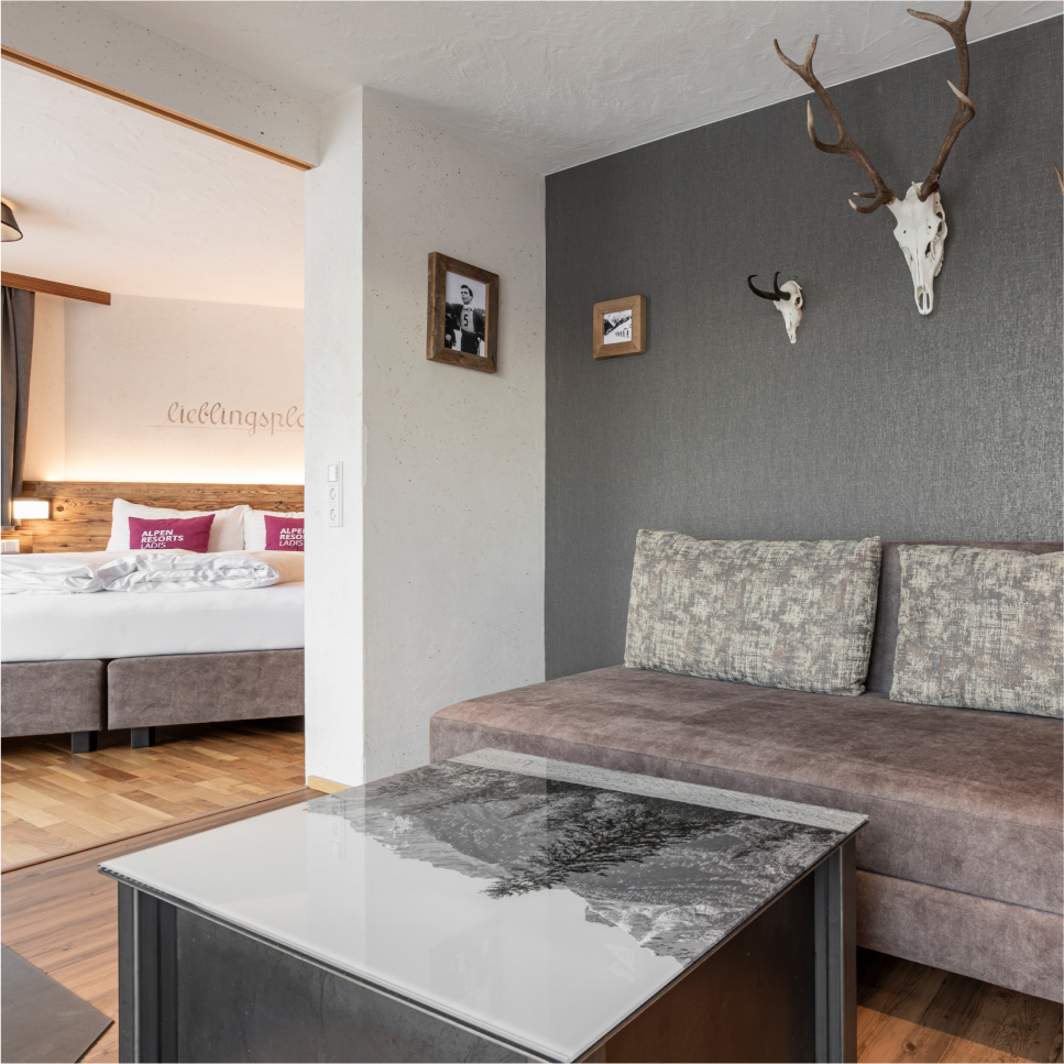 Modernes Hotelzimmer im Stil eines Familienchalets mit einem gemütlichen Bett mit weißer Bettwäsche und einem dunkel gepolsterten Kopfteil. Eine graue Akzentwand mit montierten Geweihen und gerahmten Fotos setzt den Akzent, ergänzt durch ein plüschiges graues Sofa und einen Couchtisch aus reflektierendem Glas.