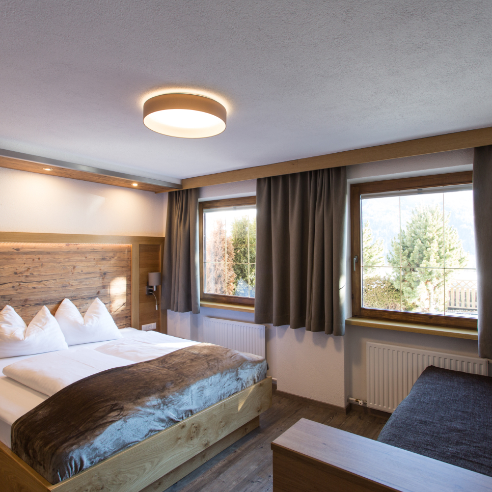 Gemütliches Hotelzimmer, das auch als gemütliche Lodge für 2-5 Personen dient, mit einem großen Bett mit weißer Bettwäsche und einer braunen Pelzdecke. Ein Kopfteil und Boden aus Holz sorgen für Wärme. Zwei Fenster mit grauen Vorhängen sorgen für natürliches Licht und einen Blick auf die Bäume, während eine moderne runde Deckenleuchte den Raum erhellt.
