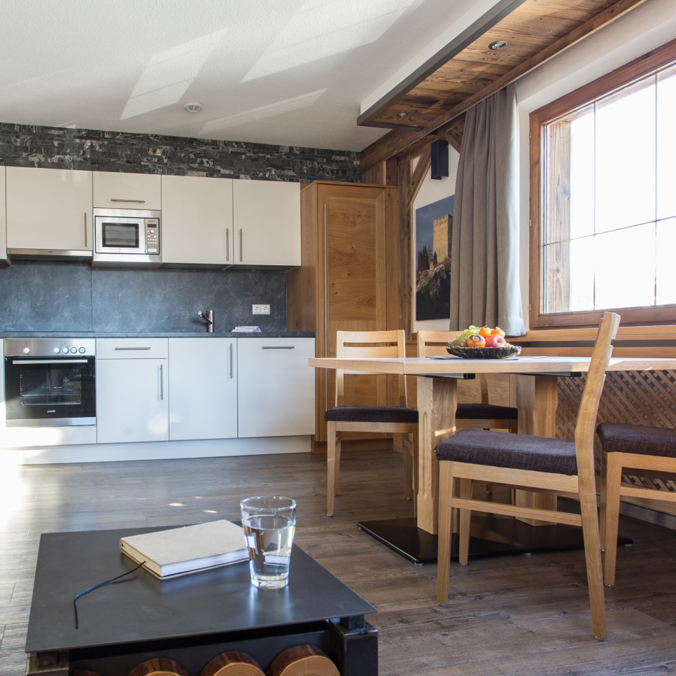Eine moderne Küche und ein Essbereich mit weißen Schränken, einem Backofen und einer Mikrowelle erwecken den Charme einer Hütte in Serfaus-Fiss-Ladis für 2-4 Personen. Ein hölzerner Esstisch mit Obst steht neben einem großen Fenster, während ein kleiner Beistelltisch über einem Weinregal ein Glas Wasser und ein Buch bereithält.