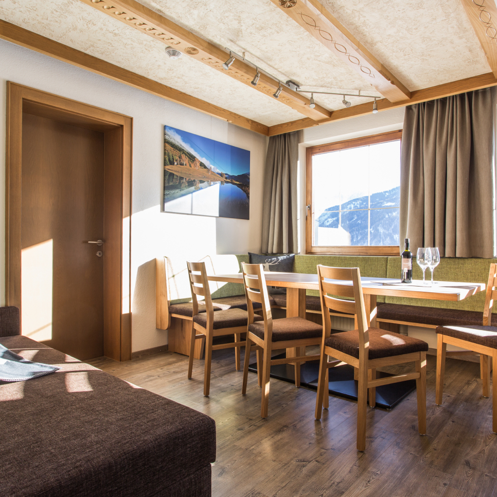 Eine gemütliche Lodge für 6-8 Personen mit Terrasse verfügt über einen Essbereich mit einem Holztisch und sechs Stühlen. Sonnenlicht strömt durch ein großes Fenster und beleuchtet ein Sofa und Wandkunst. Der Raum hat Holzböden und eine Balkendecke, und Wein und Gläser stehen einladend auf dem Tisch.