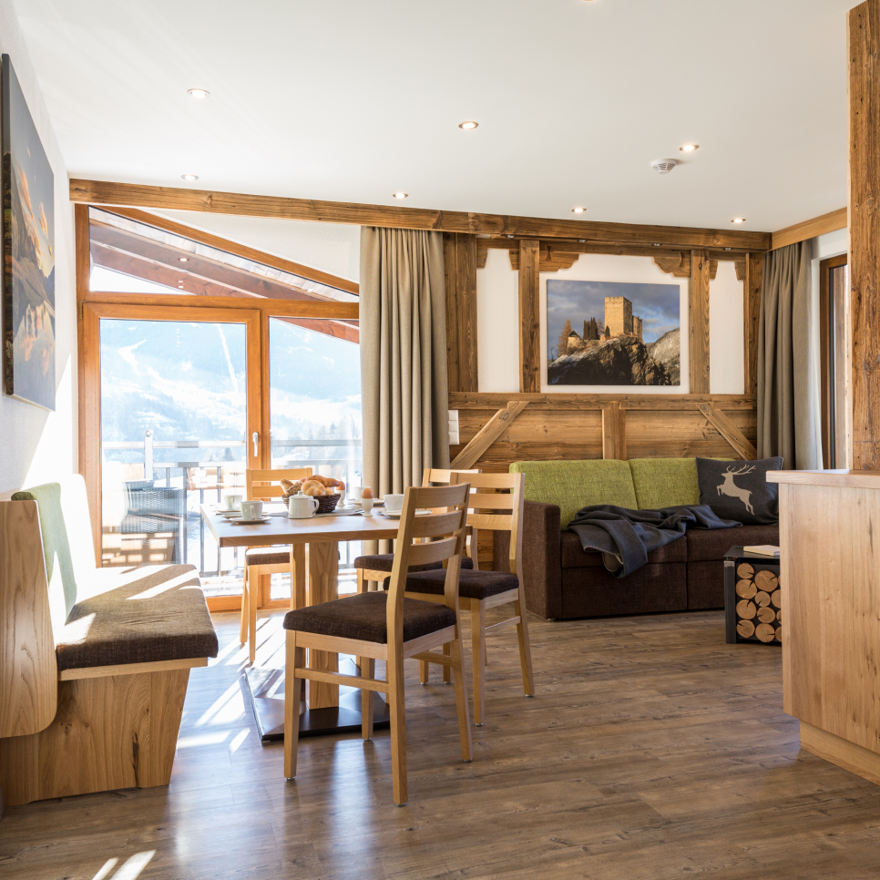 Ein gemütliches Zimmer in einer modernen Lodge für 4-6 Personen in Ladis verfügt über Holzmöbel, einen Frühstückstisch, Stühle und ein Ecksofa. Ein gerahmtes Bild schmückt die Wand. Große Fenster bieten einen malerischen Blick auf die Berge und in der Ecke liegt ein Stapel Brennholz.