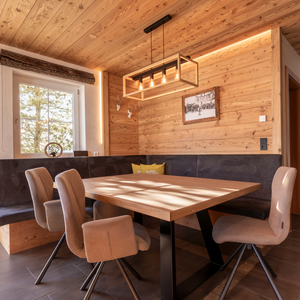 Ein gemütliches Esszimmer aus Holz in einem Luxusapartment für 6-8 Personen in Serfaus-Fiss-Ladis verfügt über einen rechteckigen Tisch, der von sechs gepolsterten Stühlen umgeben ist. Ein moderner Kronleuchter hängt darüber, während Sonnenlicht durch das Fenster fällt und die Holzwände und -decke hervorhebt, wobei ein dekoratives Wandstück sichtbar ist.