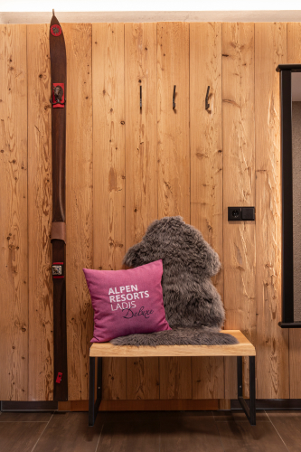 In einem gemütlichen und doch luxuriösen Ambiente besticht das hölzerne Interieur mit einem an der Wand hängenden Vintage-Ski. Darunter sorgt eine Bank mit einem pelzigen grauen Polster und einem magentafarbenen Kissen für Charme, perfekt für Gäste des Luxusapartments für 6-8 Personen in Serfaus-Fiss-Ladis. Haken bieten praktische Möglichkeiten zum Aufhängen von Gegenständen.