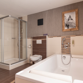 Erleben Sie die Eleganz eines modernen Badezimmers in unserem Luxusapartment für 6-8 Personen in Serfaus-Fiss-Ladis. Es verfügt über eine verglaste Dusche, eine weiße Badewanne und ein wandmontiertes WC vor Wänden mit Fliesenmuster und dunklen, holzähnlichen Bodenfliesen. Ein Bild mit einer Berglandschaft verleiht der Wanne eine ruhige Note.