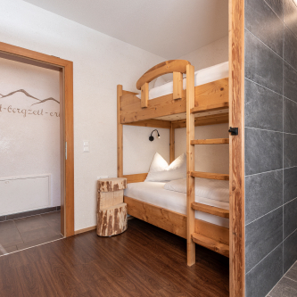 Ein gemütliches Zimmer in diesem Luxusapartment für 6-8 Personen in Serfaus-Fiss-Ladis verfügt über ein Etagenbett aus Holz mit weißer Bettwäsche. Neben dem Bett steht ein rustikaler Holzhocker. Das Zimmer verfügt über einen Holzboden und eine grau geflieste Wand, während eine offene Tür zu einem mit Kunstwerken geschmückten Flur führt.