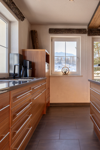Eine moderne Küche in einem Luxusapartment für 6-8 Personen in Serfaus-Fiss-Ladis mit Holzschränken und glatten Arbeitsflächen. Eine Kaffeemaschine steht inmitten schicker Einrichtung, mit einem Fenster, das den Blick auf eine verschneite Berglandschaft freigibt. Rustikale Holzbalken verleihen diesem alpinen Rückzugsort Charme.