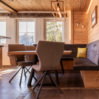 Ein gemütliches Esszimmer in einem Luxusapartment für 6-8 Personen in Serfaus-Fiss-Ladis mit Holzeinrichtung. Ein moderner Tisch ist von gepolsterten Stühlen und einer Bank umgeben. Sonnenlicht strömt durch große Fenster mit Blick auf den verschneiten Wald, während eine geometrische Hängelampe das warme Ambiente verstärkt.