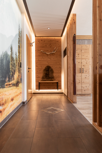 Ein Flur in einem Luxusapartment für 6-8 Personen in Serfaus-Fiss-Ladis verfügt über Holzwände und -böden, eine pelzbezogene Bank mit montierten Geweihen und ein Wandgemälde einer Waldlandschaft. Der Raum wird mit sanftem, stimmungsvollem Licht beleuchtet.