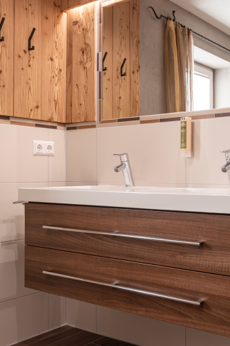 Dieses Luxusapartment für 6-8 Personen in Serfaus-Fiss-Ladis verfügt über ein modernes Badezimmer mit einem Holzwaschtisch mit zwei Schubladen, einer weißen Ablagefläche und einem Spiegel, flankiert von Holzschränken. Ein mit einem Vorhang versehenes Fenster durchflutet den Raum mit natürlichem Licht.