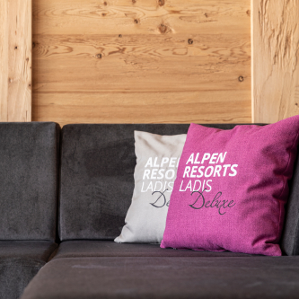 Eine gemütliche Ecke in einem Luxusapartment für 6-8 Personen in Serfaus-Fiss-Ladis besticht durch eine dunkelgraue Couch mit zwei zauberhaften Kissen. Eines ist rosa, das andere hellgrau, beide mit dem Schriftzug „ALPEN RESORTS LADIS Deluxe“ geschmückt. Die Hintergrundwand ist mit eleganten hellen Holzpaneelen versehen.