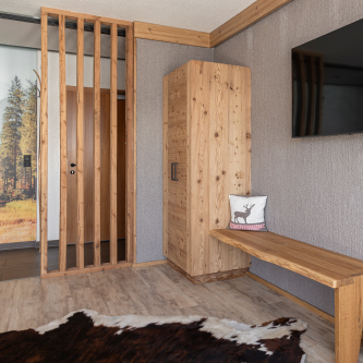 Ein rustikales Luxusapartment für 6-8 Personen in Serfaus-Fiss-Ladis verfügt über Holzelemente, darunter einen Kleiderschrank, eine Bank und einen Wandteiler. Ein Flachbildfernseher ist an einer strukturierten Wand montiert. Auf der Bank liegt ein dekoratives Kissen mit einer Hirschsilhouette, während ein Kuhfellteppich einen Teil des Holzbodens bedeckt.