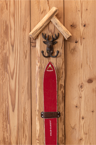 An einer dekorativen Holzwand im Luxusapartment für 6-8 Personen in Serfaus-Fiss-Ladis ist ein vertikal montierter roter Ski angebracht. Darüber befindet sich in einer kleinen hölzernen Dachkonstruktion ein Metallhaken in Form eines Hirschkopfes, der rustikale und wintersportliche Themen miteinander verbindet.