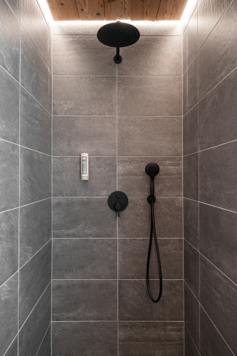Eine moderne Dusche im Luxusapartment für 6-8 Personen in Serfaus-Fiss-Ladis verfügt über grau geflieste Wände mit einem schwarzen Regenduschkopf und Handbrause. Die Holzdecke verfügt über Einbauleuchten, die eine warme Atmosphäre schaffen, die perfekt zum Entspannen einlädt.