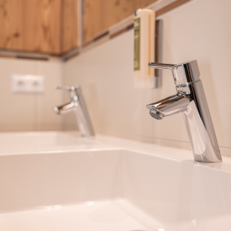 Nahaufnahme eines modernen Waschbeckens in einem Luxusapartment für 6-8 Personen in Serfaus-Fiss-Ladis mit einem eleganten Chromhahn. Im Hintergrund betonen ein weiterer Wasserhahn, ein wandmontierter Seifenspender und eine Holzverkleidung das Design. Der Fokus liegt weiterhin auf dem eleganten Wasserhahn und Waschbecken an der Vorderseite.