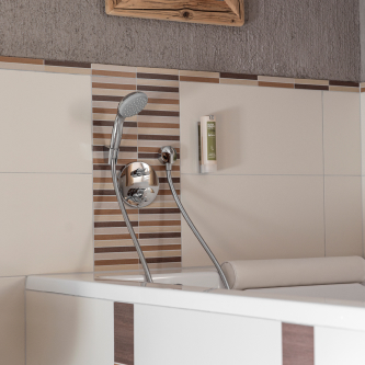 Ein modernes Badezimmer in einem Luxusapartment für 6-8 Personen in Serfaus-Fiss-Ladis mit einer Badewanne mit montiertem Handbrausekopf. Die Wand zeigt horizontale Streifen aus braunen und beigen Fliesen, und auf dem eingebauten Regal über der Wanne steht eine kleine grüne Flasche.