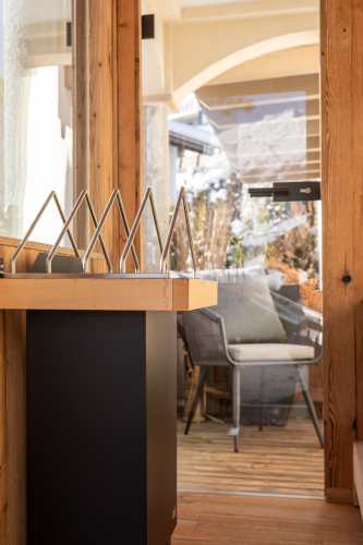 Eine Glastür mit Holzrahmen öffnet sich zu einer gemütlichen Außenterrasse mit einem Stuhl, perfekt zum Entspannen im Luxusapartment für 6-8 Personen in Serfaus-Fiss-Ladis. Im Inneren ergänzt ein moderner Briefkasten mit abgewinkelten Metallstangen das Design, da das Sonnenlicht weiche Schatten auf die Holzoberflächen wirft.