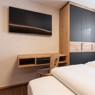 Luxusapartment für 6-8 Personen in Serfaus-Fiss-Ladis: Ein minimalistisches Schlafzimmer mit einem montierten Fernseher, einem Schreibtisch und Stuhl aus Holz sowie einem großen Kleiderschrank mit eleganter schwarzer und hölzerner Verkleidung. Das Bett mit frischer weißer Bettwäsche ist teilweise im Vordergrund sichtbar.