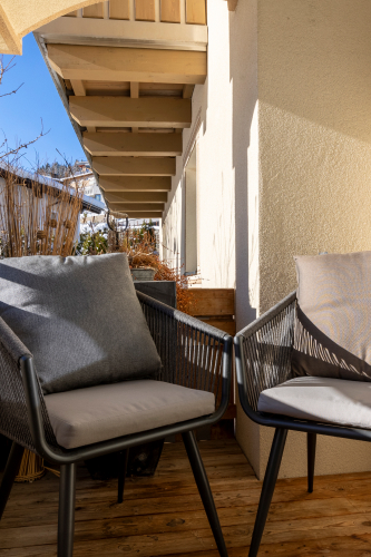 Zwei gepolsterte Stühle auf einem Holzbalkon mit beiger Wand bieten Ruhe in diesem Luxusapartment für 6-8 Personen in Serfaus-Fiss-Ladis. Ein Dachüberstand spendet Schatten, da das Sonnenlicht sanfte Schatten wirft. Blauer Himmel und üppige Pflanzen verstärken die heitere Atmosphäre.