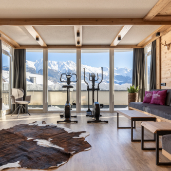 Modernes Zimmer in einem Luxusapartment für 6-8 Personen in Serfaus-Fiss-Ladis mit zwei Heimtrainern vor raumhohen Fenstern mit Blick auf die schneebedeckten Berge. Der Raum umfasst einen Kuhfellteppich, eine Holzbank, ein graues Sofa mit violetten Kissen und sanfte Beleuchtung, die natürliche Holzakzente hervorhebt.