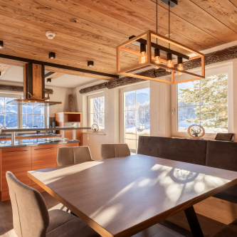 Ein gemütlicher Essbereich in einem Luxusapartment für 6-8 Personen in Serfaus-Fiss-Ladis verfügt über einen Holztisch und Stühle. Große Fenster rahmen den Blick auf die schneebedeckten Berge ein, während die Holzdecke und die moderne Hängelampe für Wärme sorgen. Im Hintergrund sind helle Küchenschränke aus Holz zu sehen.
