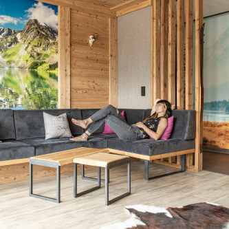 Eine Person sitzt auf einem grauen L-förmigen Sofa mit rosafarbenen Akzentkissen in einem gemütlichen Zimmer mit Holzwänden. In diesem luxuriösen Apartment für 6-8 Personen in Serfaus-Fiss-Ladis schmückt ein malerisches Berg- und See-Wandbild die Wand, während zwei kleine Holztische auf einem Kuhfellteppich auf dem hellen Holzboden stehen.