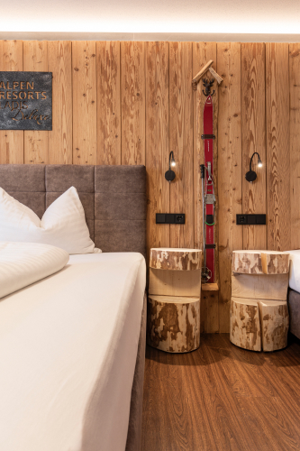 Ein gemütliches Schlafzimmer in unserem Luxusapartment für 6-8 Personen in Serfaus-Fiss-Ladis ist im Holzstil gehalten und verfügt über ein Doppelbett und weiße Bettwäsche. Nachttische aus Holzstümpfen stehen neben dem Bett, während Vintage-Skidekor die Wände schmückt. Wandleuchten sorgen für warmes Licht und verstärken den alpinen Charme.