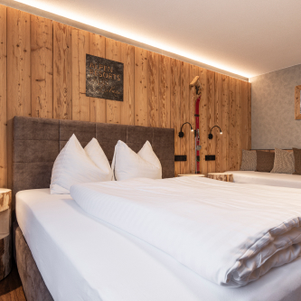 Ein gemütliches Hotelzimmer, das an ein Luxusapartment für 6-8 Personen in Serfaus-Fiss-Ladis erinnert, verfügt über ein großes Bett mit weißer Bettwäsche vor einer Akzentwand aus Holz. Sanftes Licht sorgt für Wärme, während ein kleiner Beistelltisch mit einer Lampe die behagliche Atmosphäre verstärkt. Ein Teil eines weiteren Betts oder Sitzbereichs ist sichtbar.