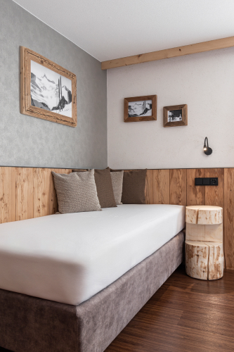 Dieses moderne Schlafzimmer, Teil eines Luxusapartments für 6-8 Personen in Serfaus-Fiss-Ladis, verfügt über ein Einzelbett mit grauer und weißer Bettwäsche, drei weiche Kissen, elegante Holzwandvertäfelungen, gerahmte Kunstwerke und zwei Holzhocker im Blockhausstil. Das Zimmer verfügt über Holzböden und ein Dekor in gedämpften Erdtönen.
