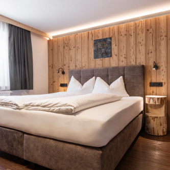 Gemütliches Hotelzimmer mit großem Bett, gepolstertem Kopfteil und weißer Bettwäsche. Die rustikale Holzwand ist dezent beleuchtet und mit Kunstwerken geschmückt. Perfekt für alle, die die Privatsphäre eines Einzelzimmers einem Luxusapartment für 6-8 Personen in Serfaus-Fiss-Ladis vorziehen. Ein Fenster ist teilweise mit Vorhängen bedeckt.