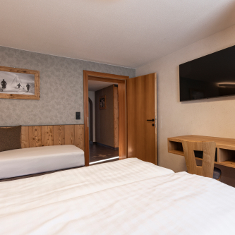 Ein gemütliches Hotelzimmer mit einem Doppelbett im Vordergrund, einem an der Wand montierten Fernseher und einem Beistelltisch aus Holz. Die Einrichtung umfasst Holzakzente und ein Wandfoto. Eine offene Tür führt in den Flur dieses Luxusapartments für 6-8 Personen in Serfaus-Fiss-Ladis.