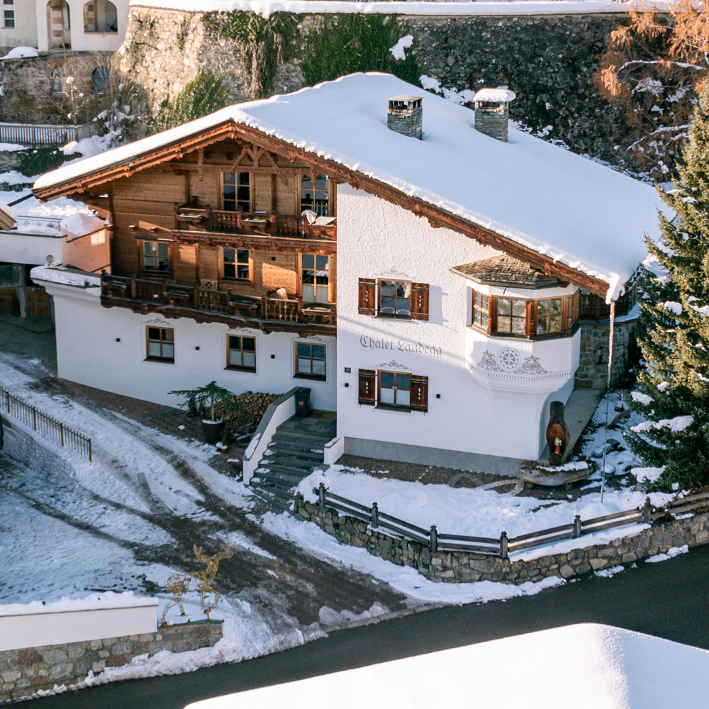 Ein malerisches Haus im Chalet-Stil in einer verschneiten Landschaft, das an das Chalet Laudegg in Ladis erinnert. Die Holzkonstruktion verfügt über einen Balkon, ein schneebedecktes Dach und charmante architektonische Details. Ein Weg führt zum Eingang, umgeben von schneebedeckten Bäumen und einem Steinzaun.