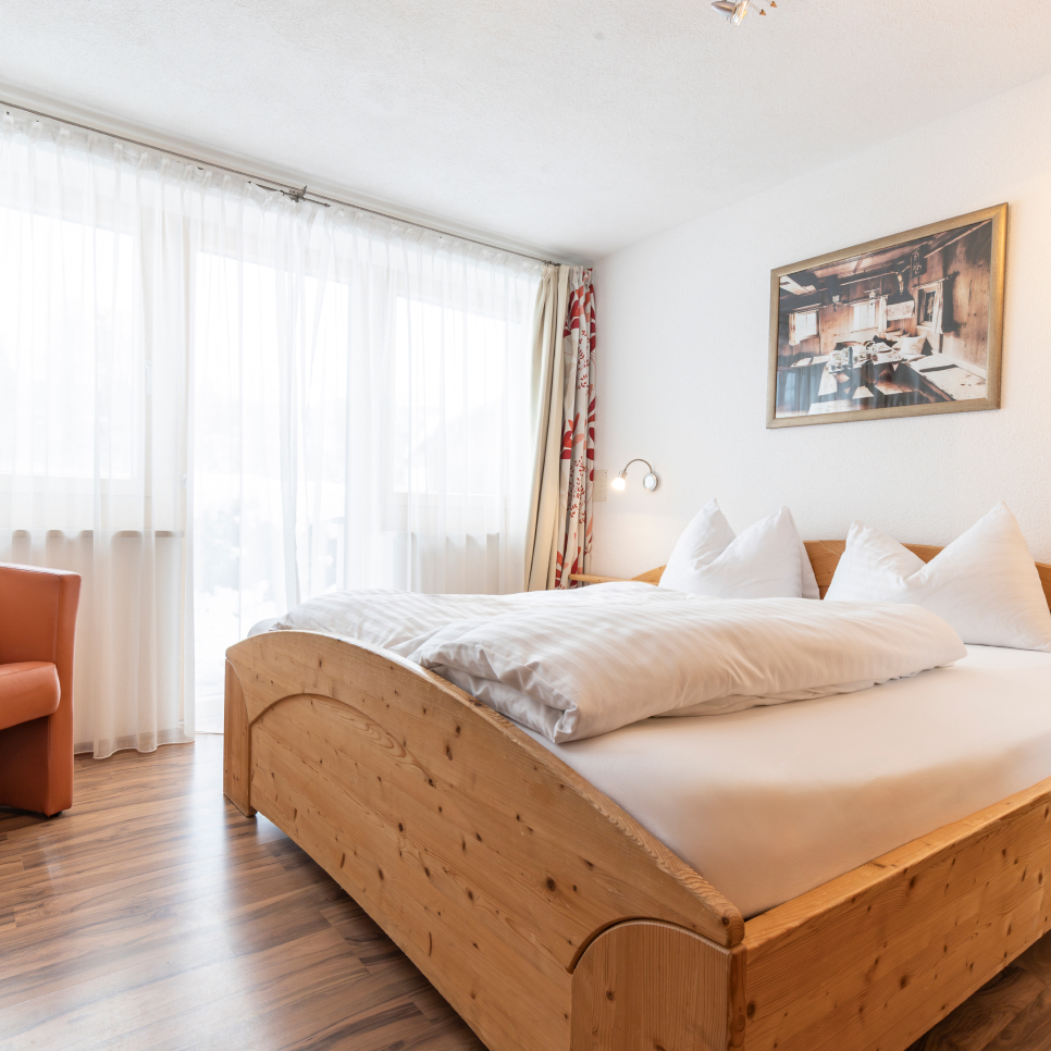 Ein gemütliches Schlafzimmer mit einem großen Holzbett mit weißer Bettwäsche und Kissen. An der weißen Wand über dem Bett hängt ein Bild. Hohe Fenster mit durchsichtigen weißen Vorhängen lassen natürliches Licht in den Raum. In der Ecke steht ein brauner Sessel.