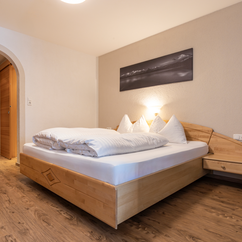 Ein gemütliches Schlafzimmer im Apartment Edelweiss für 4 Personen verfügt über ein Doppelbett aus Holz mit ordentlich angeordneten weißen Kissen und Bettwäsche. Ein sanftes Licht einer Wandlampe erhellt den Raum, während darüber ein schwarz-weißes Landschaftsfoto hängt, das den warm getönten Holzboden ergänzt.