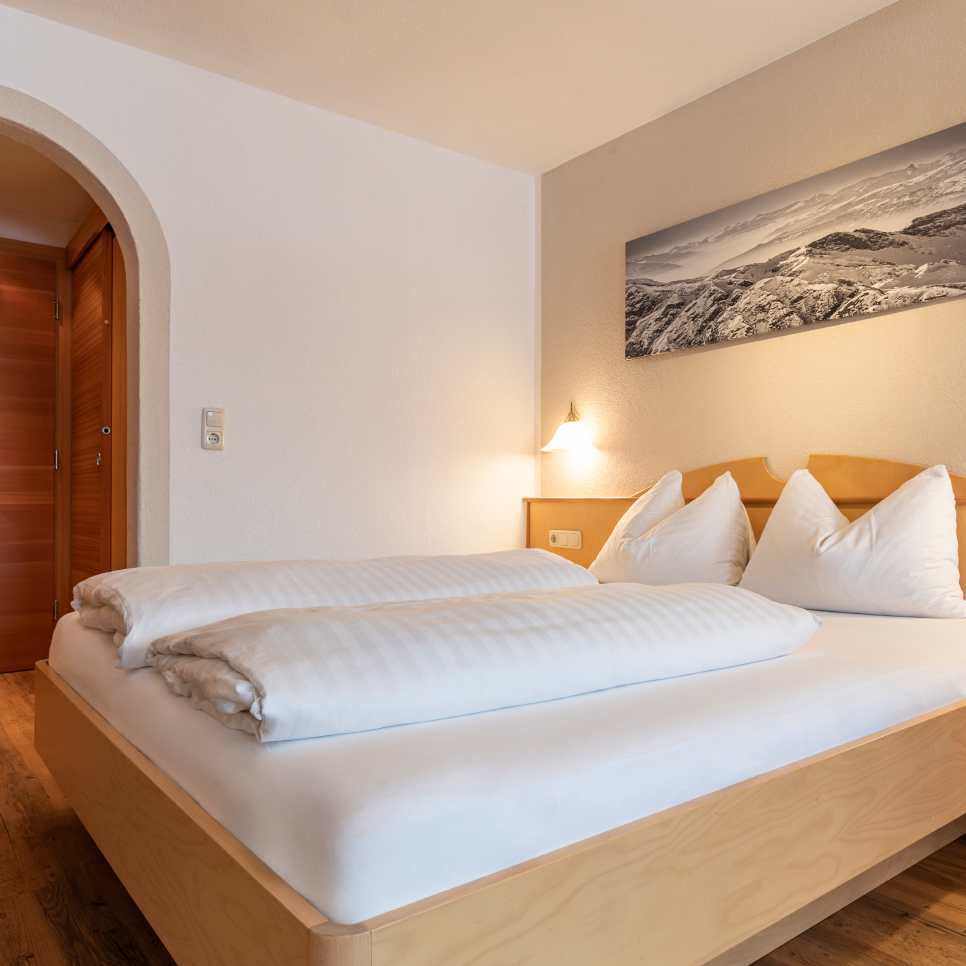 Ein gemütliches Schlafzimmer in einem Apartment für 2-3 Personen in Serfaus-Fiss-Ladis verfügt über ein Doppelbett aus Holz, weiße Bettwäsche und ordentlich angeordnete Kissen. Über dem Bett hängt ein schwarz-weißes Bergfoto, das von einer Wandlampe neben einer gewölbten Tür in warmes Licht getaucht wird.