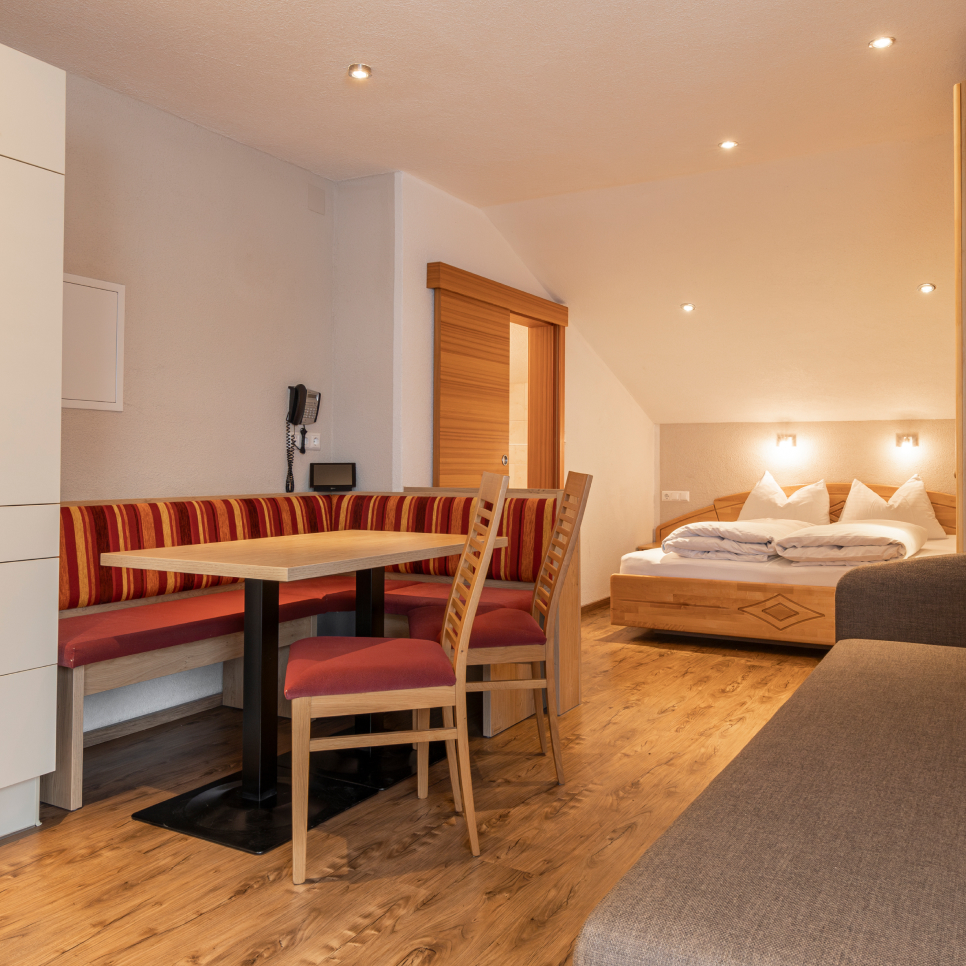Gemütliches Zimmer in einem Appartement für 2-3 Personen in Serfaus-Fiss-Ladis, ausgestattet mit Holzboden und Doppelbett mit weißer Bettwäsche. Zwei Wandleuchten beleuchten einen kleinen Essbereich mit Tisch, zwei Holzstühlen und einer Eckbank mit rot gestreiftem Polster. Eine Holzschiebetür ist sichtbar.