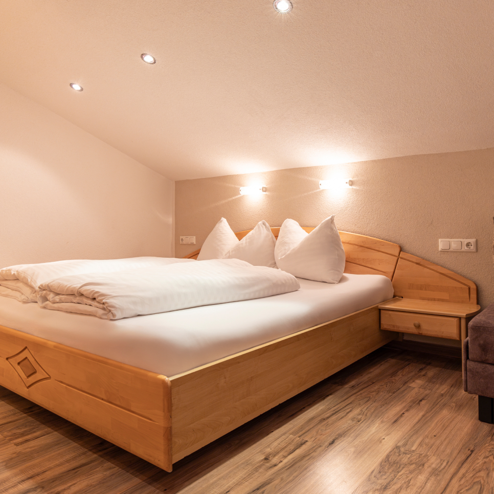 Gemütliches Schlafzimmer mit einem Doppelbett aus Holz unter einer Dachschräge. Das Bett ist hübsch gemacht, mit weißer Bettwäsche und Kissen. Warmes Licht von Wandleuchten verstärkt die einladende Atmosphäre des Zimmers. Der Boden ist aus Holz und ein kleiner Nachttisch ist sichtbar.