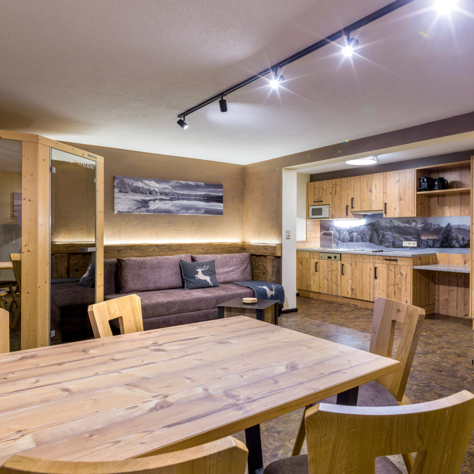 Ein gemütliches Apartment in Ladis bietet ein warmes Wohnzimmer mit einem Esstisch und Stühlen aus Holz. Eine Couch mit Kissen mit Hirschmotiven steht an der Wand. Die Küche mit Holzschränken und einem aufgehängten Landschaftsfoto vervollständigt diese charmante Umgebung für 2-5 Personen im Apartment 4.