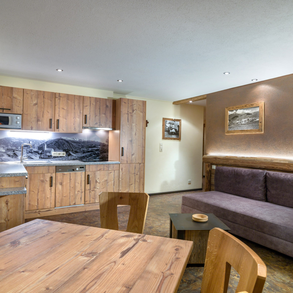 Gemütliche Küchenzeile mit holzgetäfelten Schränken und malerischen Kunstwerken. Im Apartment 5 für 4-5 Personen in Ladis bietet ein kleiner Essbereich Holzstühle und einen Tisch. Ein elegantes Sofa unter einem Bilderrahmen verstärkt die einladende Atmosphäre.