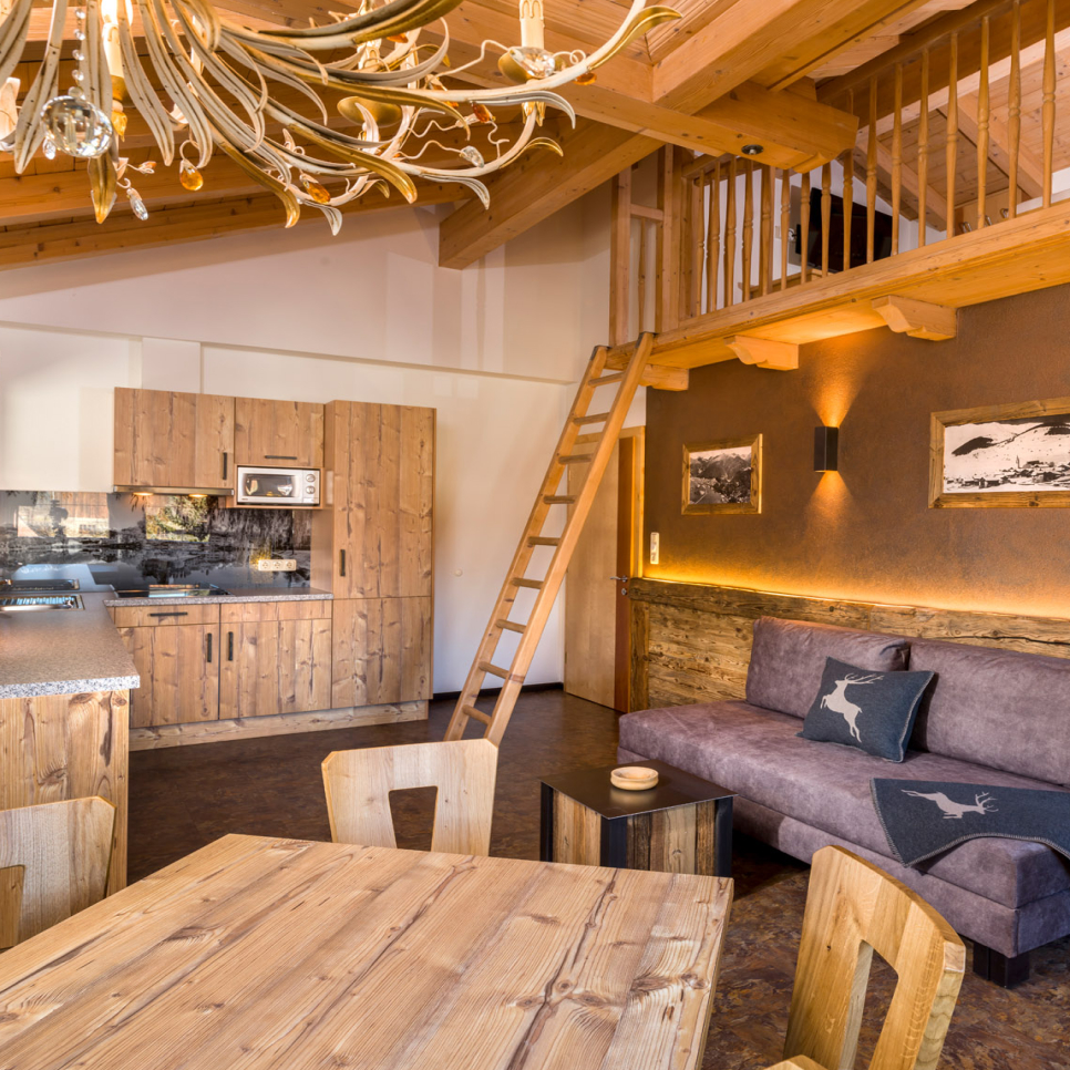 Rustikales Wohnzimmer in einem großen Apartment für 6–8 Personen in Serfaus-Fiss-Ladis mit Holzdecke und -möbeln. Eine Leiter führt zu einem gemütlichen Loft. Sofa mit Kissen mit Hirschmuster und strukturierte braune Wand mit gerahmten Bildern schaffen ein warmes Ambiente, ergänzt durch Kronleuchter und Essbereich.